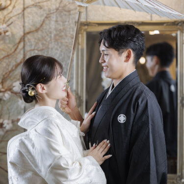 白い和傘の下で新郎の胸に手を添え、見つめ合う白無垢の花嫁。心斎橋店のスタジオで撮影した和装フォトウェディング
