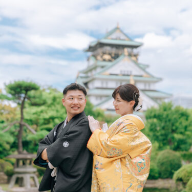 大阪城の天守を背に、新郎の肩に手を添えて寄り添う色打掛の花嫁。日本庭園の松が映える和装フォトウェディング