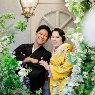 ランプとアーチ窓の前で腕を組む紋付袴の新郎と、その肩に手を添える色打掛の花嫁。笑顔あふれる和装スタジオフォト