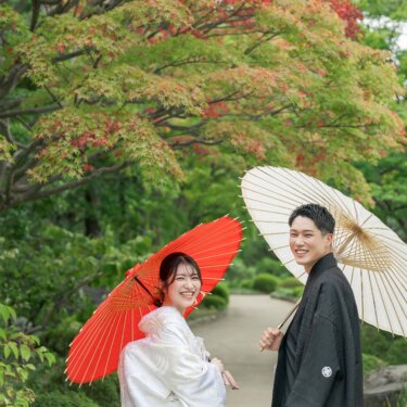 赤く色づいたモミジを背に、和傘を持って振り返り笑い合う新郎新婦。夏の日本庭園で撮る和装前撮り