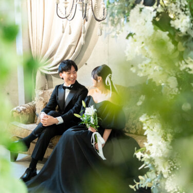 緑の葉越しに、ソファで向き合って笑う新郎と黒のカラードレスの花嫁をのぞき見るように撮った前撮り写真。前ボケが自然な雰囲気のスタジオの一枚