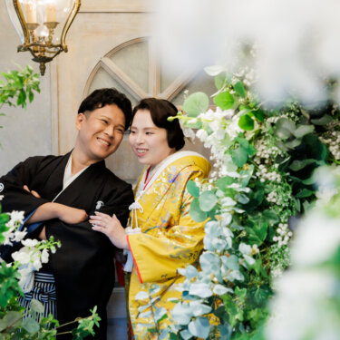 白い花とグリーンの装花越しに、肩を寄せて笑う紋付袴の新郎と色打掛の花嫁。心斎橋店のスタジオで撮る和装前撮り