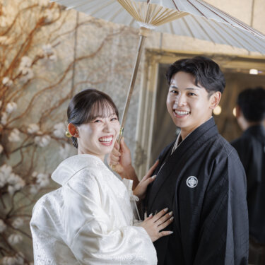 ドライフラワーの枝と白い和傘の下で、こちらを振り返って笑う新郎新婦の和装スタジオフォト