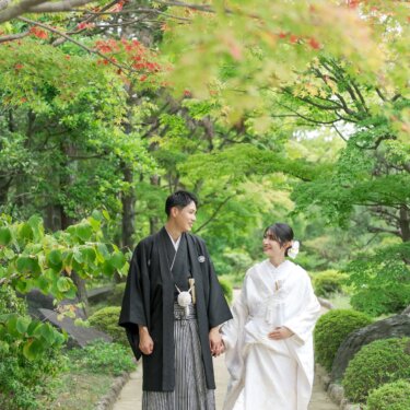 モミジと緑に囲まれた庭園の小道を手をつないで歩く、紋付袴の新郎と白無垢の花嫁。大仙公園で撮る和装前撮り
