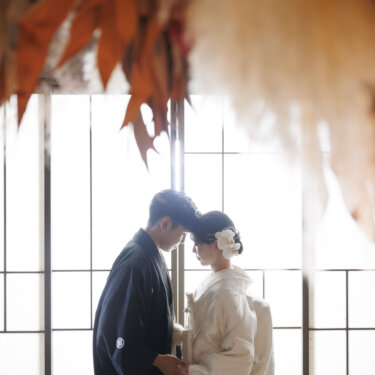 ドライフラワー越しに、窓辺でおでこを寄せて手をつなぐ紋付袴の新郎と白無垢の花嫁。大阪での和装前撮り