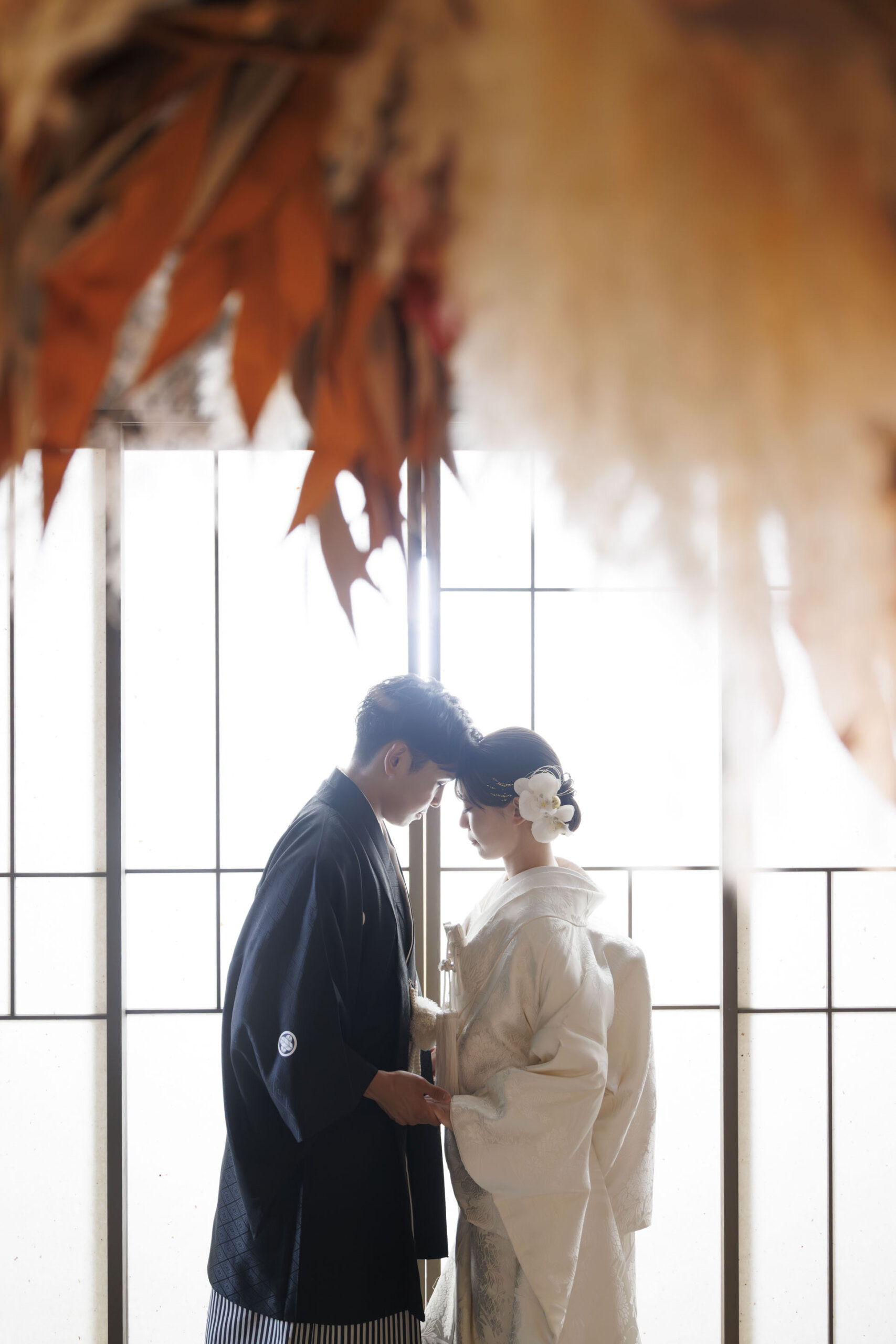 障子の前で額を寄せ合う羽織袴の花婿様と白無垢の花嫁様、ドライフラワーが美しくぼけた和室の写真
