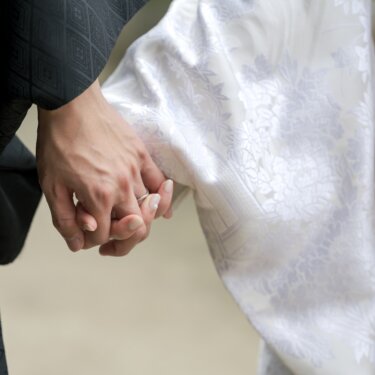 紋付の袖と白無垢の袖が重なる中で、指輪を付けた手をつなぐ新郎新婦の手元アップ。和装前撮りの定番カット