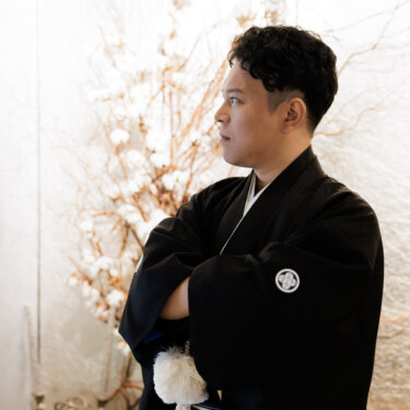 ドライフラワーの枝を背に、腕を組んで横を向く黒紋付袴の新郎。凛とした表情を写した和装前撮りの新郎ソロカット