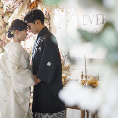 花の壁とドライフラワーで飾られた心斎橋店のスタジオで、おでこを寄せ合う新郎新婦。グリーンを前ぼけにした和装前撮り