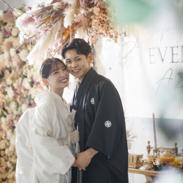 パンパスグラスとフラワーウォールの前で寄り添い、笑顔を見せる白無垢の花嫁と紋付袴の新郎。心斎橋店の和装スタジオフォト