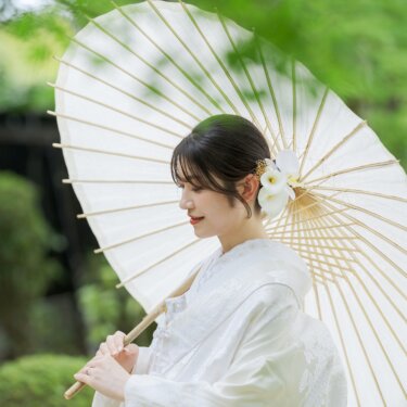 白い和傘を肩にのせ、うつむいて微笑む白無垢の花嫁。胡蝶蘭の髪飾りが映える大仙公園の新緑のロケーションフォト