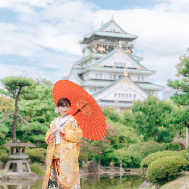 赤い和傘をさして池のほとりに立つ、黄色の色打掛の花嫁。大阪城天守閣と石灯籠が並ぶ大阪城公園の和装前撮り