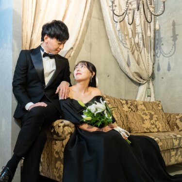 シャンデリアとカーテンのクラシカルなソファ席で、黒のカラードレスの花嫁が隣の新郎を見上げて微笑む結婚写真。黒×黒の洋装コーディネート