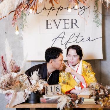 'Happily Ever After'のタペストリーとドライフラワーの前で、頬を寄せて笑う新郎新婦。日付の小物を置いた和装前撮り