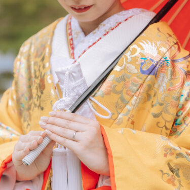 鳳凰の刺繍が入った黄色の色打掛と、赤い和傘を持つ花嫁の手元アップ。指輪が光る和装前撮りのディテールカット