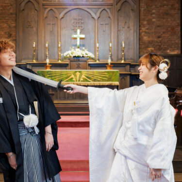 祭壇前で番傘を刀のように構えて花嫁と戯れる紋付袴の新郎と、笑顔の白無垢の花嫁。川口基督教会で撮る楽しい和装前撮りのポーズ