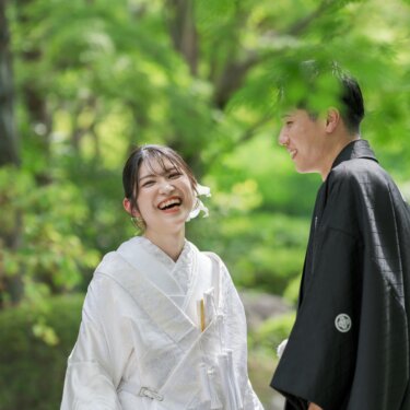 新緑の木漏れ日の中で、新郎を見上げて大笑いする白無垢の花嫁。大仙公園日本庭園で撮る笑顔の和装前撮り