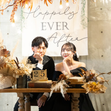 'Happily Ever After'のタペストリーとドライフラワーを飾ったテーブルで、頬杖をついて並ぶ黒いタキシードの新郎と黒のカラードレスの花嫁。大阪のスタジオで撮る洋装前撮り