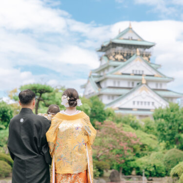 天守閣を見上げる新郎と色打掛の花嫁、抱っこされた赤ちゃんの後ろ姿。家族で叶える大阪城公園の和装前撮り