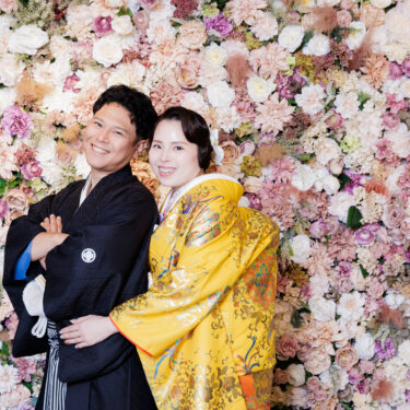 ピンクと白の花が一面に咲くフラワーウォールの前で寄り添う紋付袴の新郎と黄色の色打掛の花嫁。心斎橋のスタジオフォト