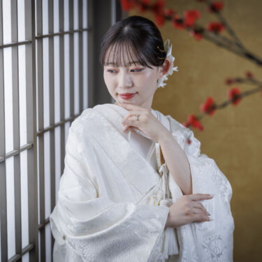 格子窓と梅の枝の前で、あごに手を添えて流し目を送る白無垢の花嫁。心斎橋店のスタジオでの和装前撮り