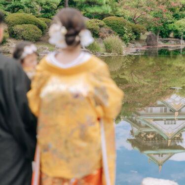 池の水面に天守閣が映るリフレクションを、新郎新婦と赤ちゃんの後ろ姿越しに写した大阪城公園の和装前撮り