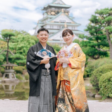 大阪城天守閣を背に、扇子を手に並んで立つ紋付袴の新郎と色打掛の花嫁。王道の構図で撮る和装の結婚写真