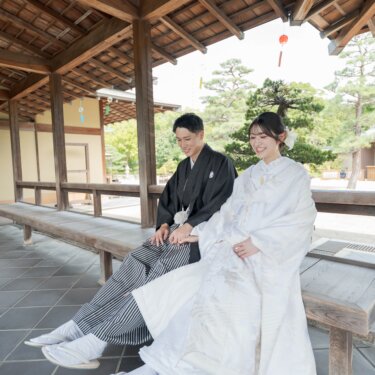 木造の回廊のベンチに並んで座り、笑い合う紋付袴の新郎と白無垢の花嫁。大仙公園日本庭園で撮る結婚写真