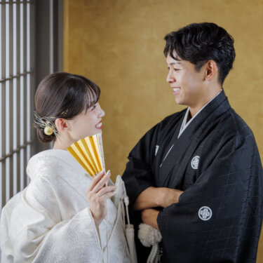 金色の壁と格子窓の前で、扇子を持つ白無垢の花嫁と腕を組んだ新郎が笑顔で見つめ合う和装フォトウェディング