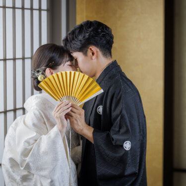 格子窓の光の中、金色の扇子で顔を隠してキスをする新郎新婦。大阪の和装スタジオフォトで人気の顔隠しポーズ
