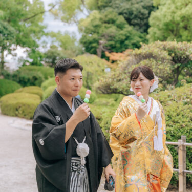 庭園の小道でお団子を手に笑い合う、紋付袴の新郎と金色の色打掛の花嫁。食べ歩き風の和装前撮り
