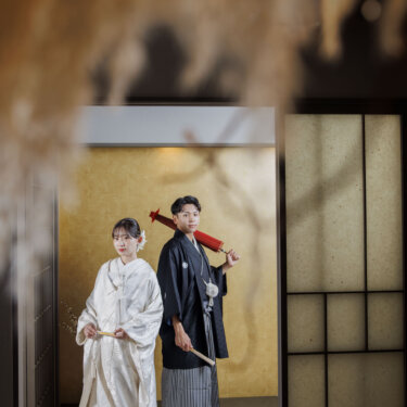 ドライフラワー越しに床の間の前で並ぶ、赤い和傘を肩にかけた新郎と白無垢の花嫁の和装前撮り