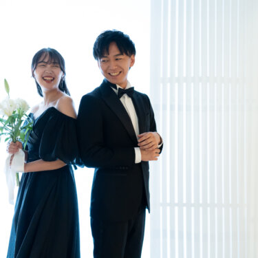 明るい窓辺で背中合わせに立ち、楽しそうに笑う黒いタキシードの新郎と黒のカラードレスの花嫁。シンプルな背景で黒×黒コーデが際立つスタジオフォト