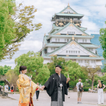 天守閣の前の広場で、お団子を手に向かい合って手をつなぐ新郎新婦。遊び心あふれる和装ロケーションフォト