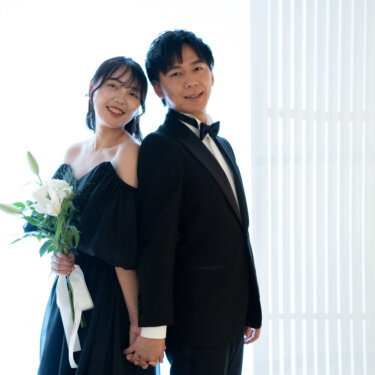白いブラインドの光を背に、背中合わせで手をつなぎ笑顔を見せる黒いタキシードの新郎とユリのブーケを持つ黒のカラードレスの花嫁。大阪のスタジオで撮る前撮り