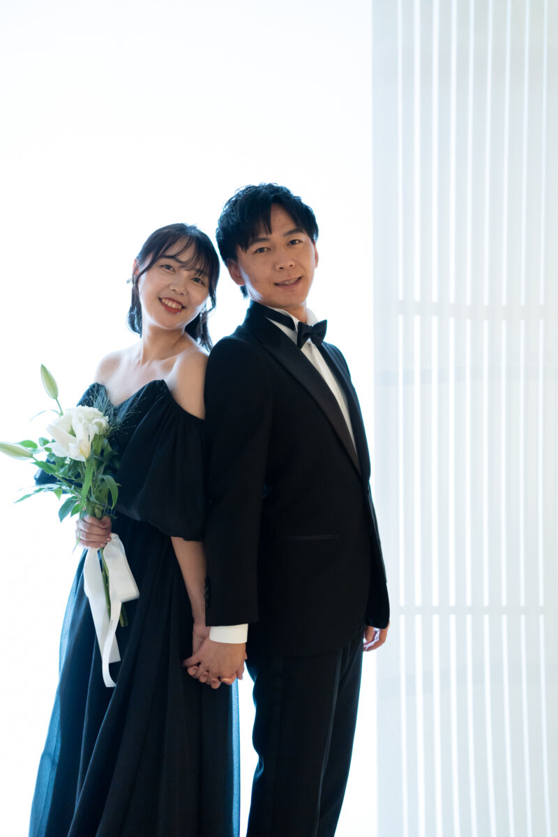 白いブラインドの光を背に、背中合わせで手をつなぎ笑顔を見せる黒いタキシードの新郎とユリのブーケを持つ黒のカラードレスの花嫁。大阪のスタジオで撮る前撮り