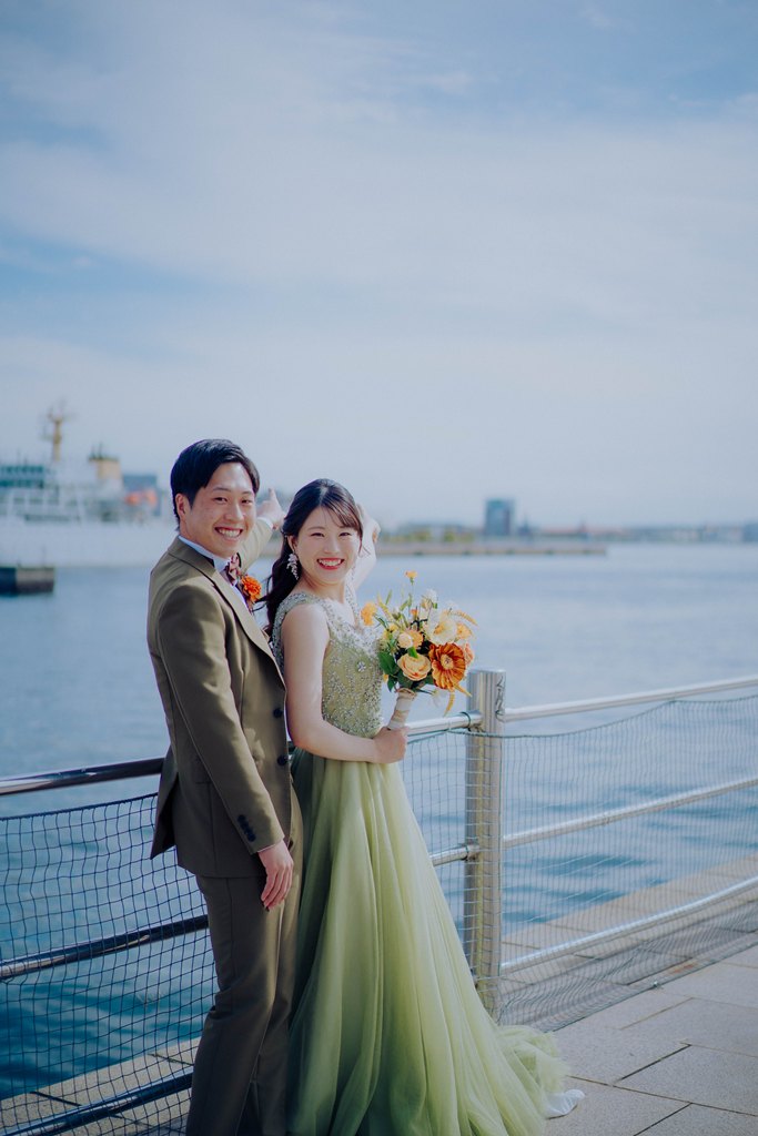 神戸ハーバーランドの海沿いのデッキで客船を背に寄り添う、グリーンのカラードレスの花嫁とオリーブのスーツの新郎の前撮り