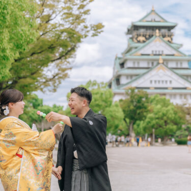 天守閣を背景に、新郎が花嫁にお団子を差し出すワンシーン。色打掛で撮る大阪城公園での楽しい和装前撮り