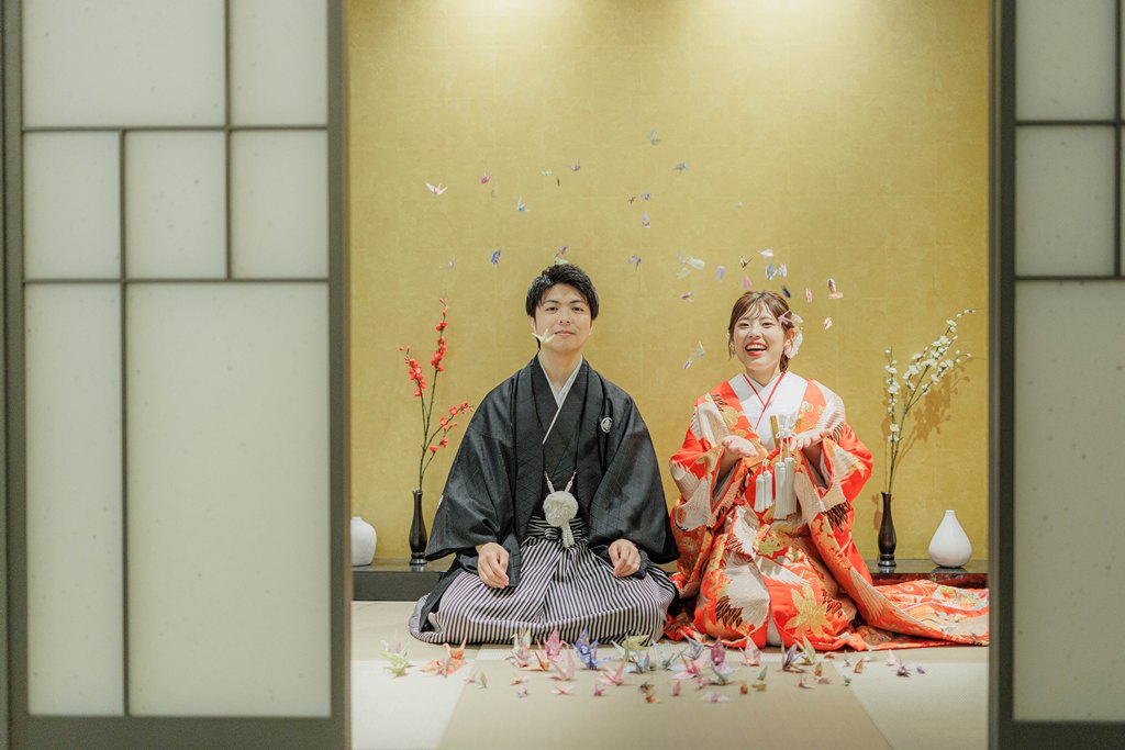 金色の壁の和室で、朱色の色打掛の花嫁様と羽織袴の花婿様が笑顔で折り鶴を舞い上げている写真