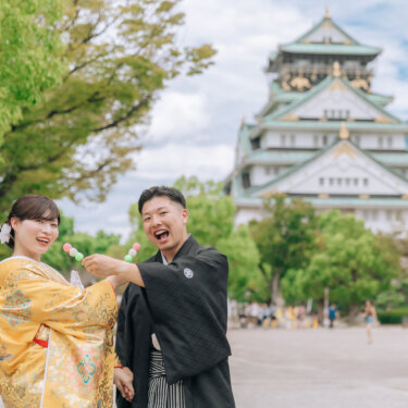 お城を背に、お団子を食べさせ合って笑う紋付袴の新郎と色打掛の花嫁。笑顔あふれる和装フォトウェディング