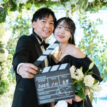 フラワーアーチの下で頬を寄せ、カチンコをカメラに向けて笑う黒いタキシードの新郎と黒のカラードレスの花嫁。遊び心のある結婚写真のポーズ