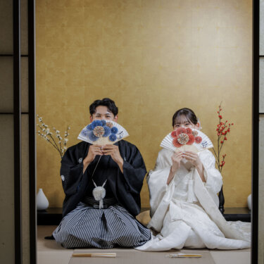 床の間の前に座り、花柄の扇子で顔を隠して笑う新郎新婦。遊び心のある顔隠しポーズの和装スタジオフォト