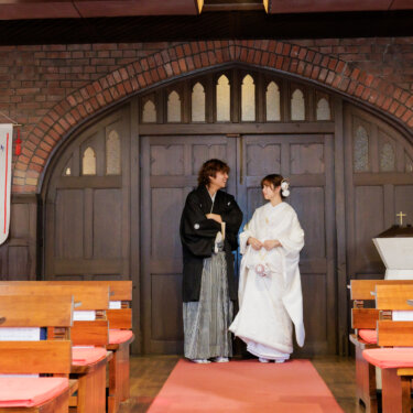 教会のバナーが下がるアーチ扉の前に並ぶ紋付袴の新郎と白無垢の花嫁。レンガ造りの川口基督教会で撮る和装フォトウェディング