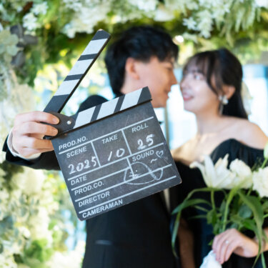 カチンコを掲げる新郎と、ユリのブーケを持って新郎を見つめる黒のカラードレスの花嫁。白い花のアーチを背にした映画風の前撮り