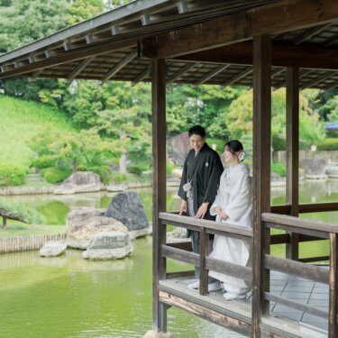 朱色の東屋が見える池の回廊に並んで立つ新郎新婦。築山と庭石が広がる大仙公園日本庭園の和装前撮り