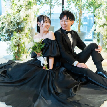 グリーンのフラワーアーチの前の床に並んで座り、黒のカラードレスの裾を大きく広げて笑い合う新郎新婦。心斎橋店のスタジオで撮る洋装前撮り