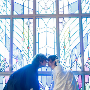 光あふれるステンドグラスの前で、手をつないでおでこを寄せ合う紋付袴の新郎と白無垢の花嫁。教会で撮るロマンチックな和装前撮り