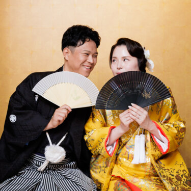 白と黒の扇子を顔の横にかざして笑い合う紋付袴の新郎と色打掛の花嫁。遊び心のある和装スタジオフォトのポーズ
