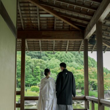 屋根付きの回廊の手すりにもたれ、池の向こうの緑を眺める新郎新婦の後ろ姿。大仙公園日本庭園での和装前撮り