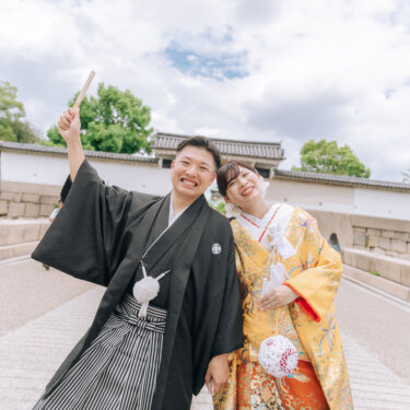 大阪城の石垣と門の前で、扇子を高く掲げておどける新郎と寄り添う色打掛の花嫁。元気いっぱいの和装前撮り